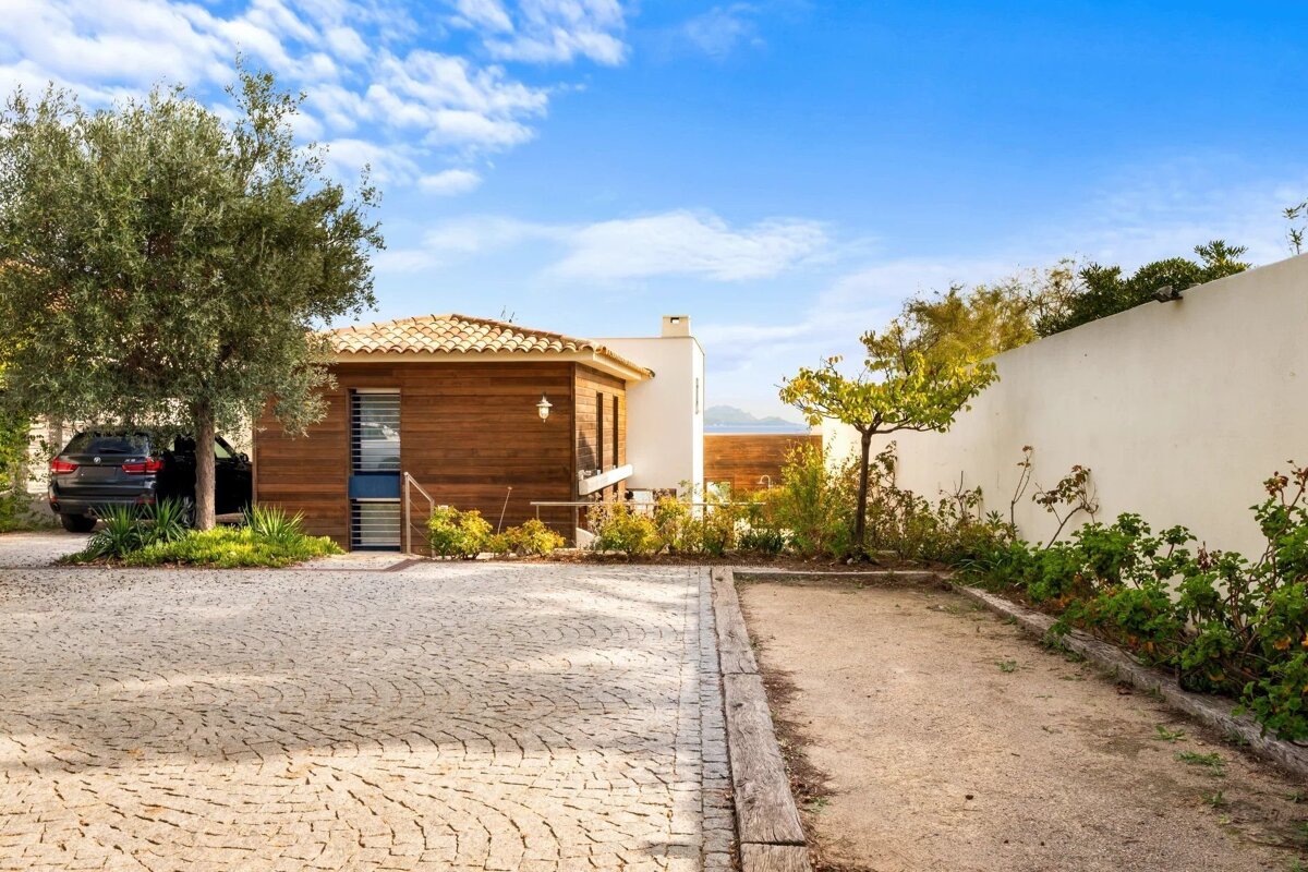 A modern wooden house with a tiled roof, paved driveway, and black car under a tree. A garden area is next to it, under a bright blue sky.