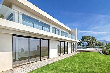 A modern white luxury villa with large glass windows, a wooden deck, and a lush green lawn under a bright blue sky.