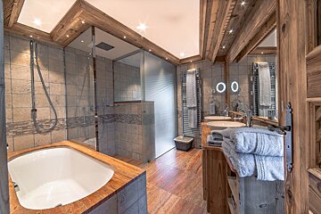 Cozy, rustic bathroom with a bathtub, glass shower, dual sinks, and warm wood accents & tiled walls.