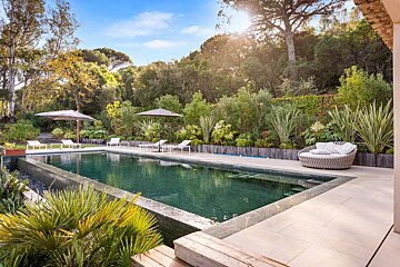 A beautiful, dark-tiled swimming pool nestled within a lush, green garden under a sunny sky. Loungers, umbrellas, and a daybed furnish the poolside.