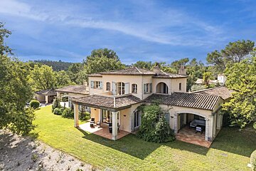 A grand, cream-colored villa with terracotta tiled roofs and blue shutters, nestled amidst lush green lawns and dense trees under a clear blue sky.