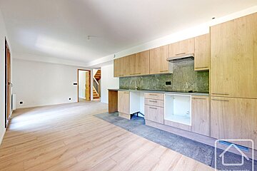 An empty, open-concept room with light wood floors. It features a new kitchen with light wood cabinets, gray counters, a stone backsplash, and appliance spaces. A staircase is visible.