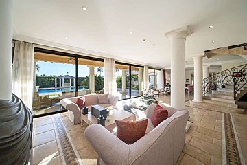 A luxurious living room with large windows overlooking a pool, featuring elegant sofas, classic columns, a grand staircase, and a Christmas tree.