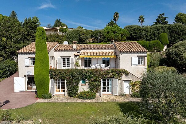 A sunny, Mediterranean-style villa with white walls, terracotta roof, yellow awning, and white shutters, nestled on a lush hillside with gardens.