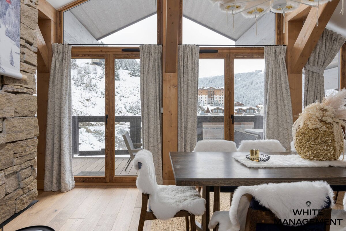 A cozy dining area in a ski chalet with large windows showcasing a snowy mountain view. Features a wooden table, fur-covered chairs, and rustic stone/wood elements.