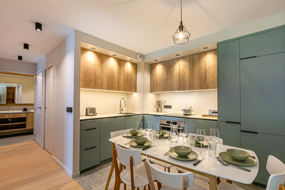 Modern kitchen with sage green base & wood upper cabinets, under-cabinet lighting, and a dining table set with dishes.