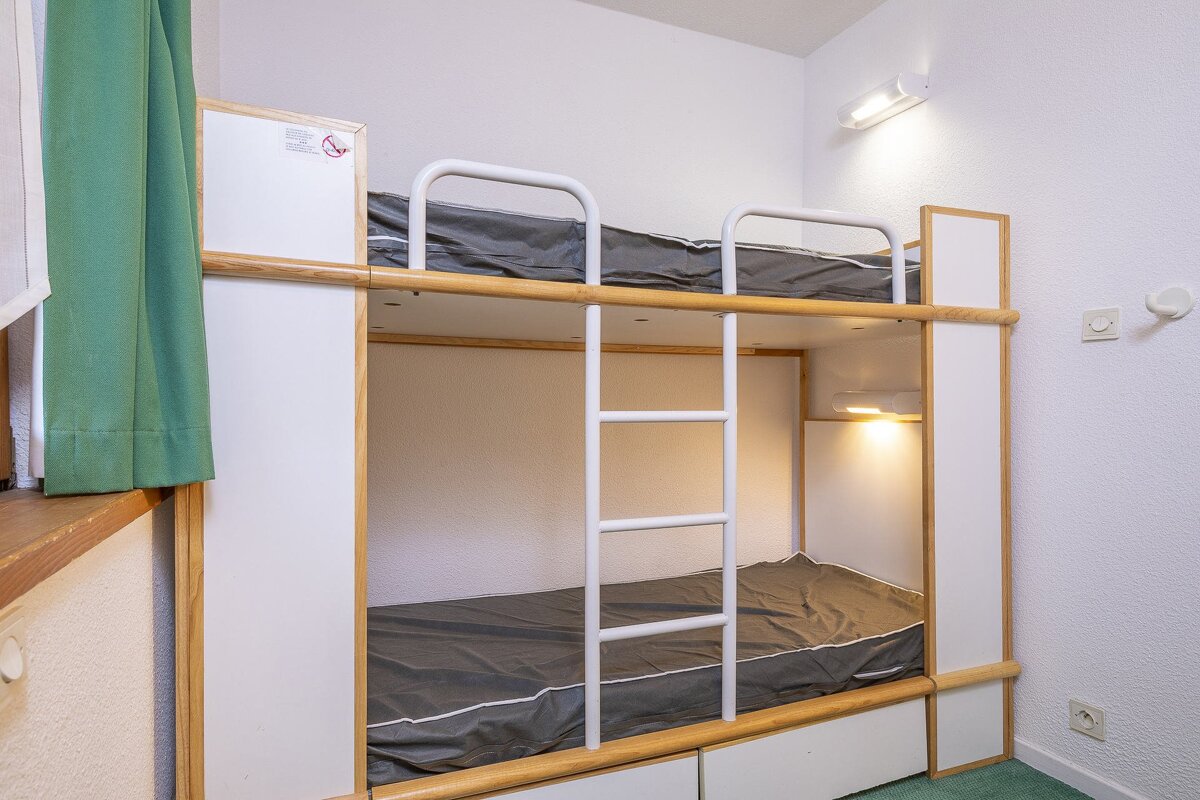 A brightly lit room features modern bunk beds with white and wood frames, grey mattresses, and a ladder. Green curtains hang by a window.