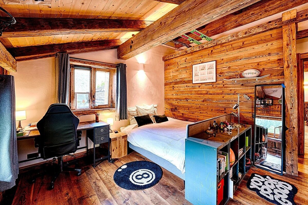 A cozy, wood-paneled bedroom with exposed ceiling beams. It includes a bed, a desk with a chair, a window, and quirky decor like an 8-ball rug.