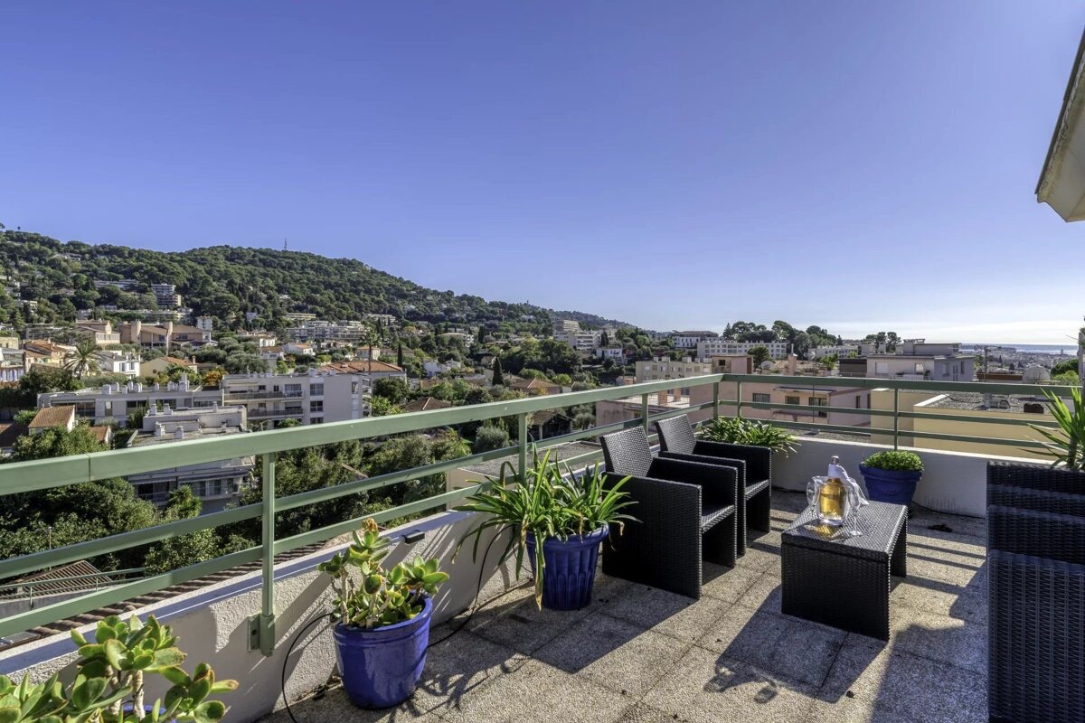 Sunny rooftop terrace with outdoor seating, potted plants, and a panoramic view of a hillside town and the distant sea under a clear blue sky.