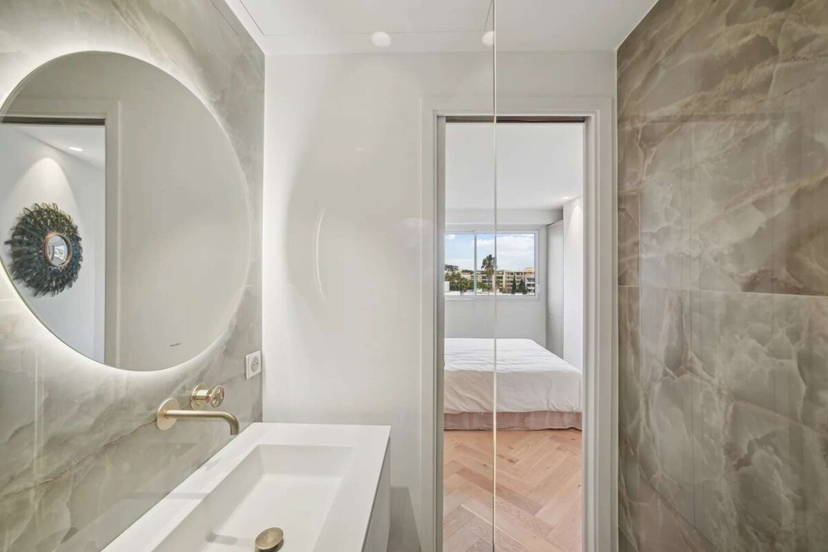 A sleek bathroom with a lighted mirror and marble walls connects to a bright bedroom via a glass partition, revealing a white bed and herringbone floor.