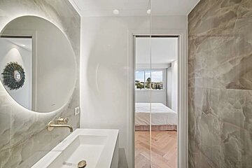 A sleek bathroom with a lighted mirror and marble walls connects to a bright bedroom via a glass partition, revealing a white bed and herringbone floor.