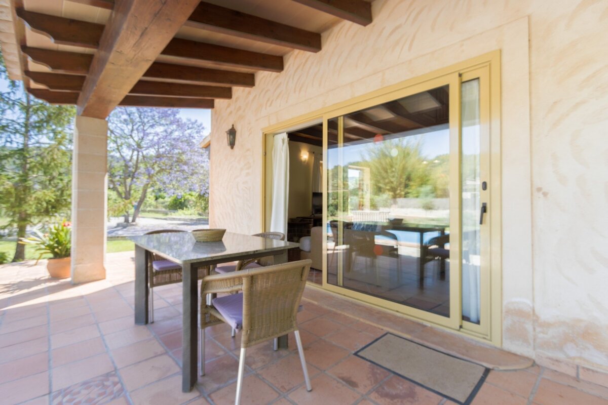 A covered patio with a dining set and terracotta floor. Sliding glass doors open to a lush garden with blooming purple trees.