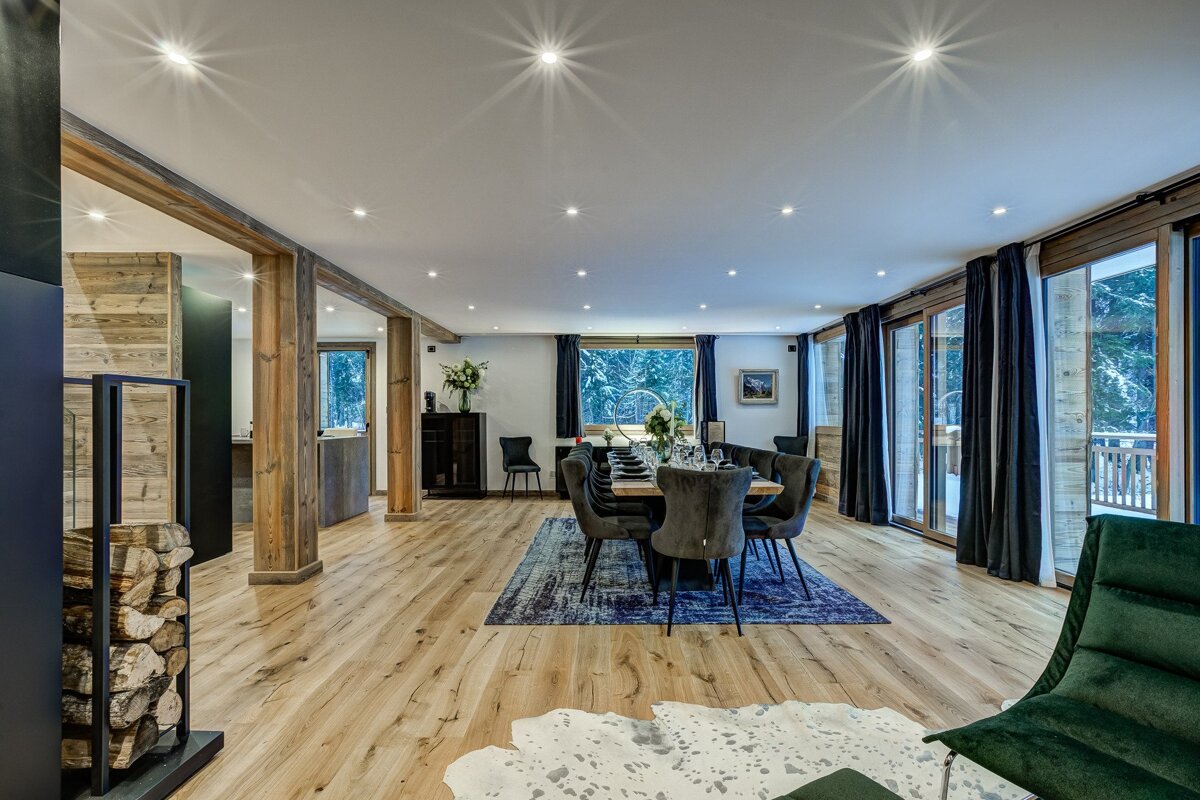 Spacious modern chalet dining room with wood floors, exposed beams, a long dining table, and large windows overlooking a snowy forest. Firewood stack visible.
