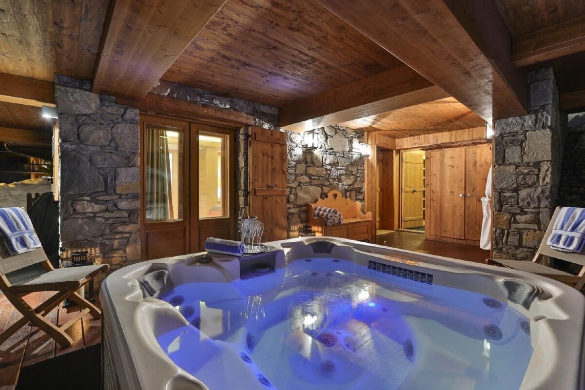 Cozy indoor hot tub with blue lights, rustic stone walls, and wooden chalet-style interior, suggesting a luxurious mountain retreat.
