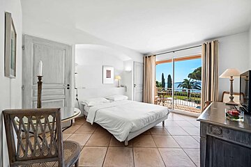 A bright white bedroom with a bed, light tile floor, and a balcony offering a stunning sea view with palm trees and a blue sky.