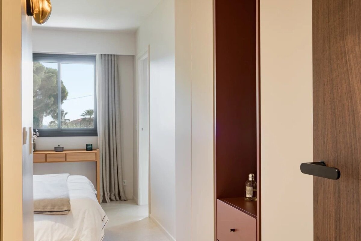 A modern bedroom featuring a white bed, a wooden desk by a window with outdoor trees, and a contrasting maroon built-in storage niche with a pink drawer.