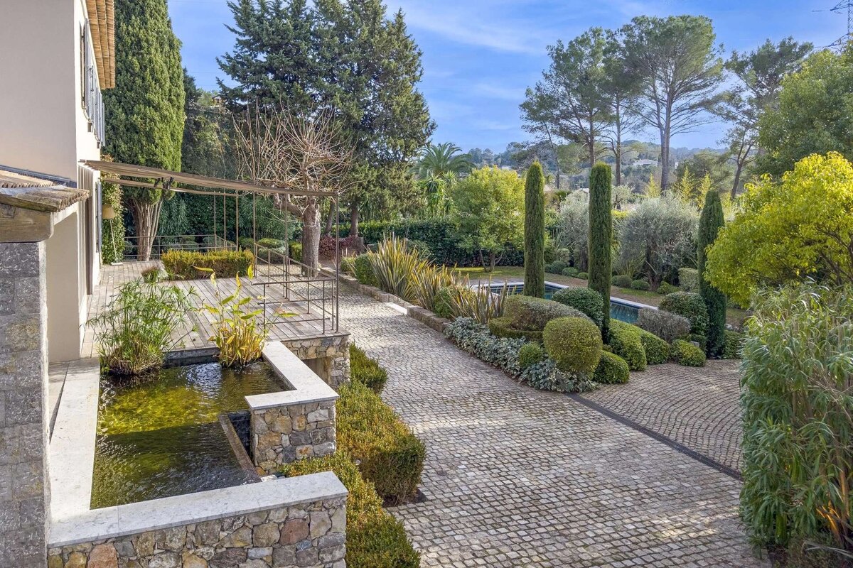 A lush, manicured garden with a paved driveway, water feature, and diverse trees next to a house under a bright blue sky.