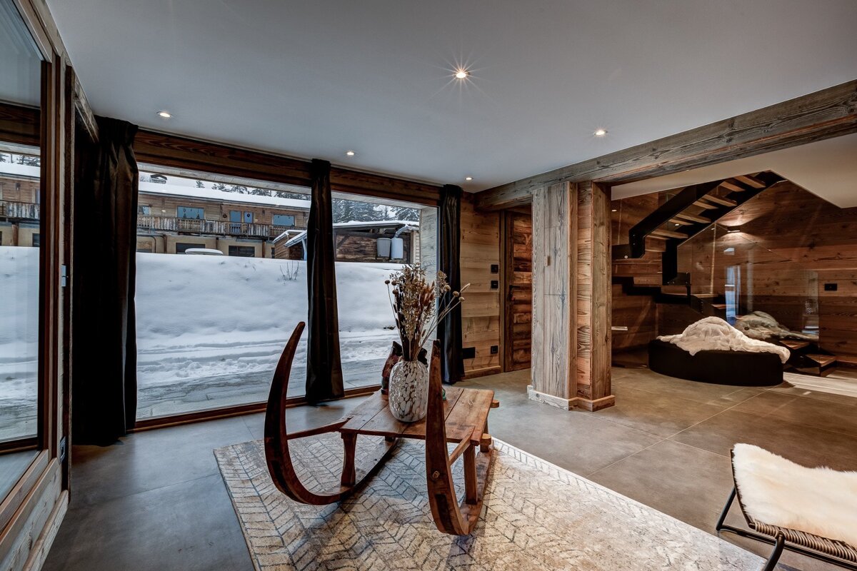A chic chalet interior with a snowy outdoor view. Rustic wood walls, concrete floors, a unique sleigh coffee table, and modern glass staircase create a cozy-yet-sleek ambiance.