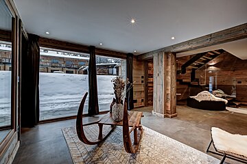 A chic chalet interior with a snowy outdoor view. Rustic wood walls, concrete floors, a unique sleigh coffee table, and modern glass staircase create a cozy-yet-sleek ambiance.