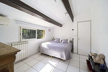 A bright, white bedroom with a bed, white tiled floor, exposed wooden beams, and a window. An AC unit and radiator are visible.