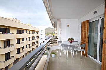 A tiled apartment balcony with a round table and chairs, potted plants, and views of neighboring buildings.