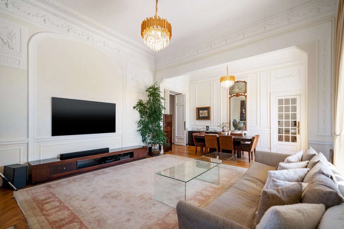 An elegant living and dining room features ornate white walls, a large TV, a cozy sofa, glass coffee table, and a dining area under golden chandeliers.