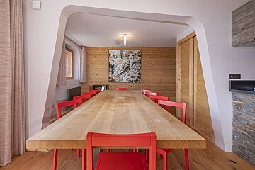 A long wooden dining table with bright red chairs, framed by a modern white archway in a room with wood-paneled walls.