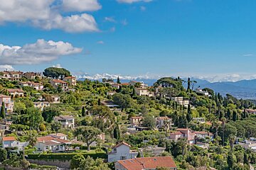 Hillside houses in France with lush greenery, under a blue sky with distant snow-capped mountains.