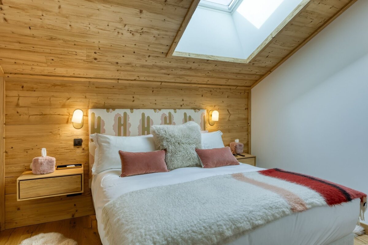 A cozy, wood-paneled bedroom with an angled ceiling and skylight, featuring a bed dressed in white, plush textures, and pink-red accents.