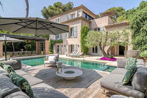 A luxurious Mediterranean villa with a sparkling pool, stylish outdoor lounging, and a shaded dining area, surrounded by lush greenery on a sunny day.