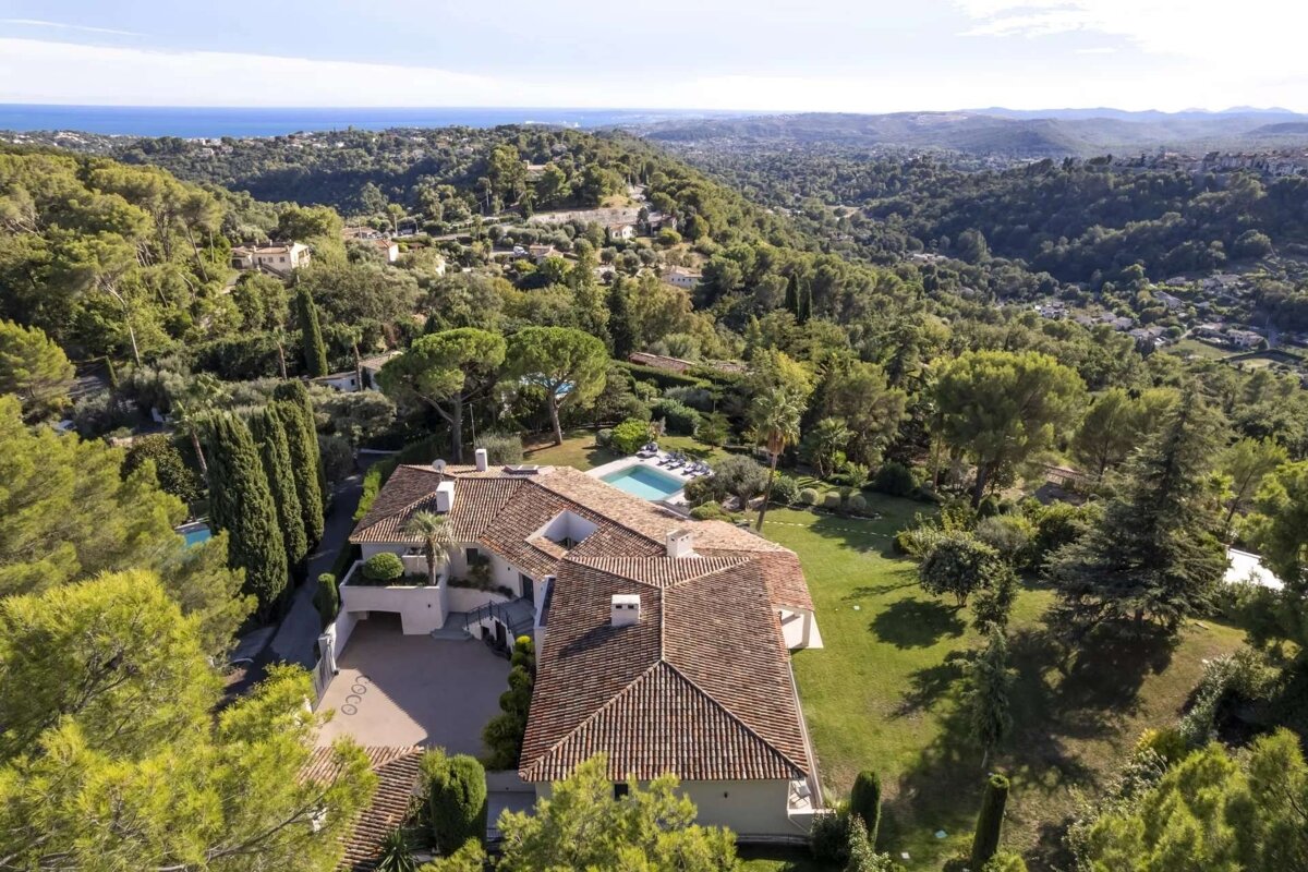 Aerial view of a large villa with a pool, surrounded by lush green hills and trees, with the distant sea visible under a clear sky.
