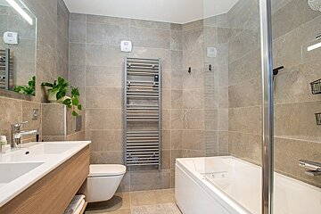 Modern bathroom with grey tiles, a wooden vanity, wall-mounted toilet, towel radiator, and bathtub with a glass screen.