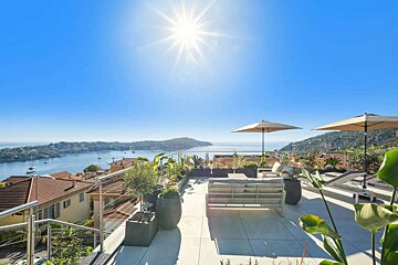 A sun-drenched rooftop terrace offers stunning bay views, featuring outdoor seating, umbrellas, and potted plants above red-tiled houses.