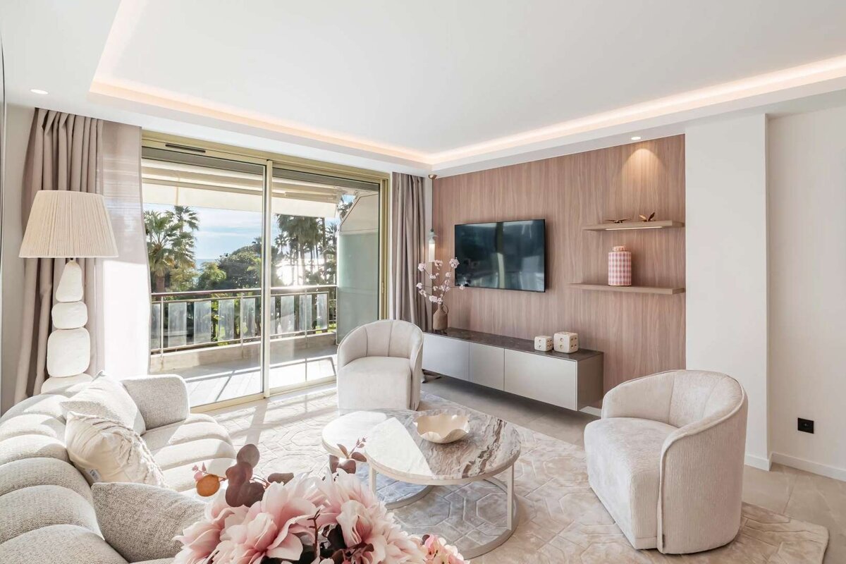 Bright, modern living room with light furniture, a wood-paneled TV wall, and a balcony with a scenic ocean and palm tree view.