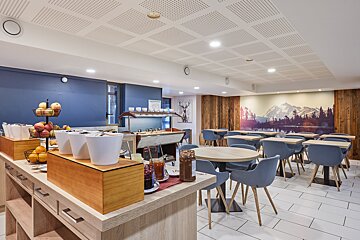 A modern hotel breakfast room featuring a buffet with fresh food, blue chairs, wooden tables, and a scenic mountain mural backdrop.