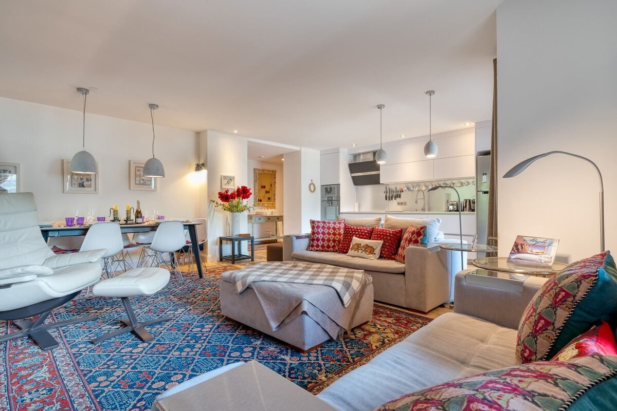 A bright, open-plan living room, dining area, and modern kitchen. It features colorful patterned rugs, cozy sofas, and an elegant dining table.