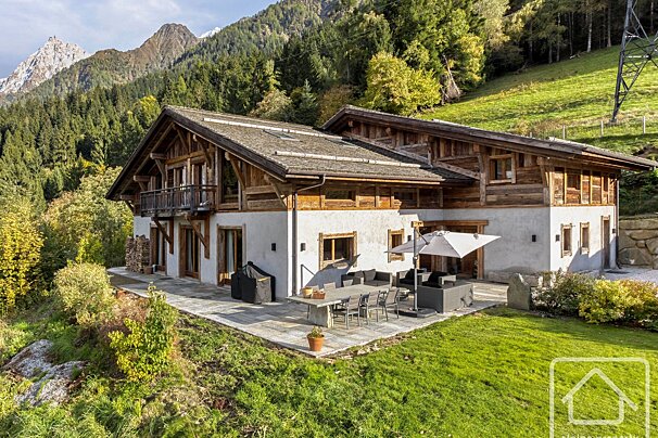 A rustic wooden and white chalet sits amidst forested mountains. It features a large patio with outdoor furniture, a sun umbrella, and a green lawn.