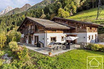 A rustic mountain chalet with a large outdoor patio, seating, and umbrella, surrounded by green lawns and forested mountains under a clear sky.