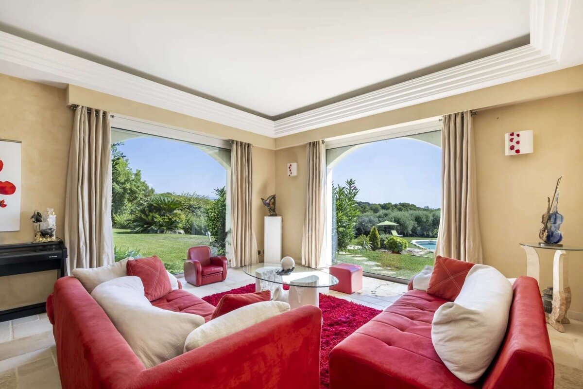 A luxurious living room featuring vibrant red sofas, a matching rug, and expansive arched windows that open to a lush green garden with a pool.