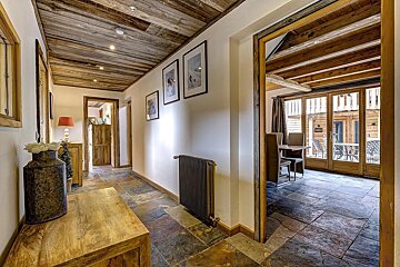 Cozy chalet interior with wood ceilings, white walls, and slate floors. A hallway with art leads to a dining room with large glass doors.