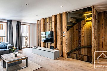 A modern living room features prominent wood paneling and beams, a wall-mounted TV, and a built-in staircase. A sofa, coffee table, and large windows create a warm, inviting space.