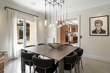 Modern dining room with a square table, black chairs, a multi-bulb light fixture, and a framed portrait of David Bowie on the wall.