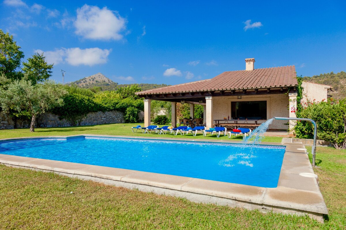 A beautiful villa with a large swimming pool, surrounded by green grass and sun loungers. Mountains rise in the background under a clear blue sky.