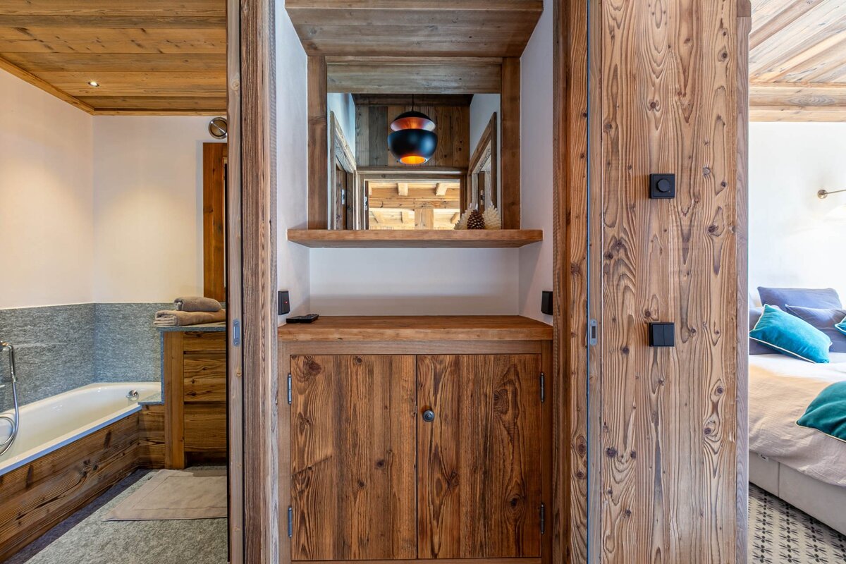 Rustic wooden interior with a mirror, shelf, and cabinet. Views of a bathroom with a tub on the left and a bedroom on the right.