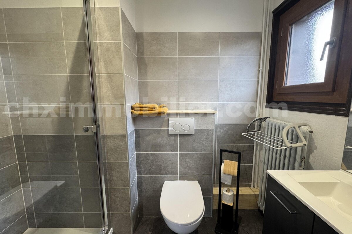 Modern bathroom with gray tiles, featuring a walk-in shower, toilet, sink, window, and radiator.