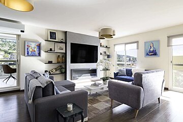 A bright, modern living room featuring grey sofas, a large TV above a linear fireplace, built-in shelving, and a balcony with a scenic view.