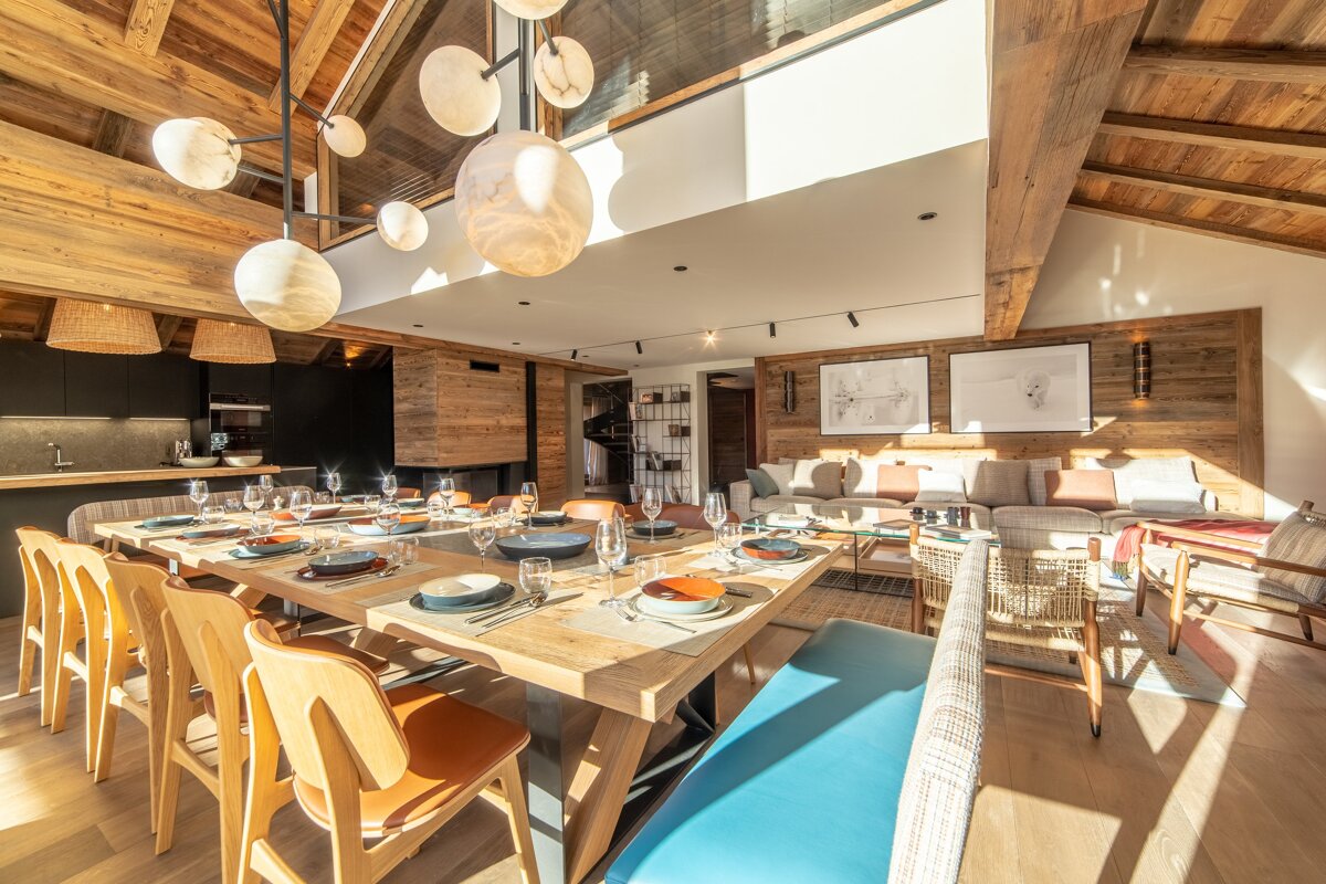 Modern chalet interior with an open-plan dining and living area. Features a vaulted wooden ceiling, a large set table, and warm natural light.