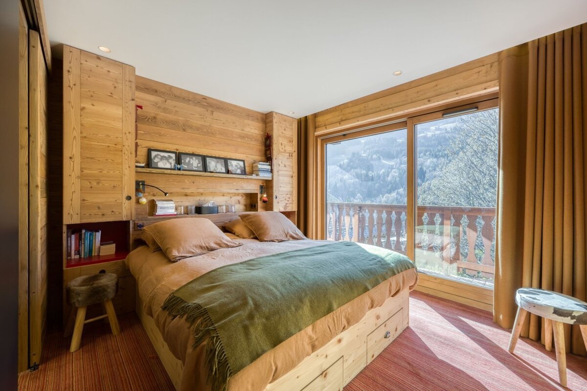 A cozy, wood-paneled chalet bedroom features a large bed with earthy bedding, built-in shelving, and a stunning mountain view from a full-length window.