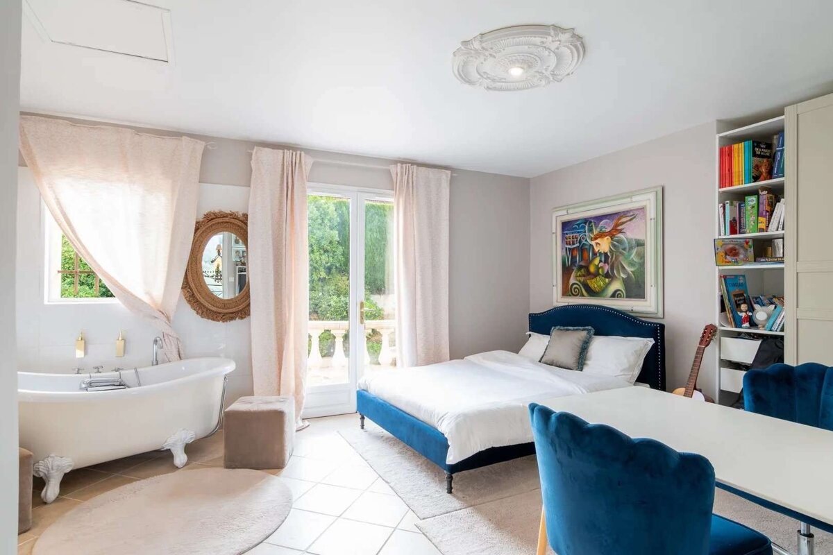 Bright room with a blue bed, white clawfoot tub, and French doors leading to a balcony. Features include pink curtains, a bookshelf, and a colorful painting.