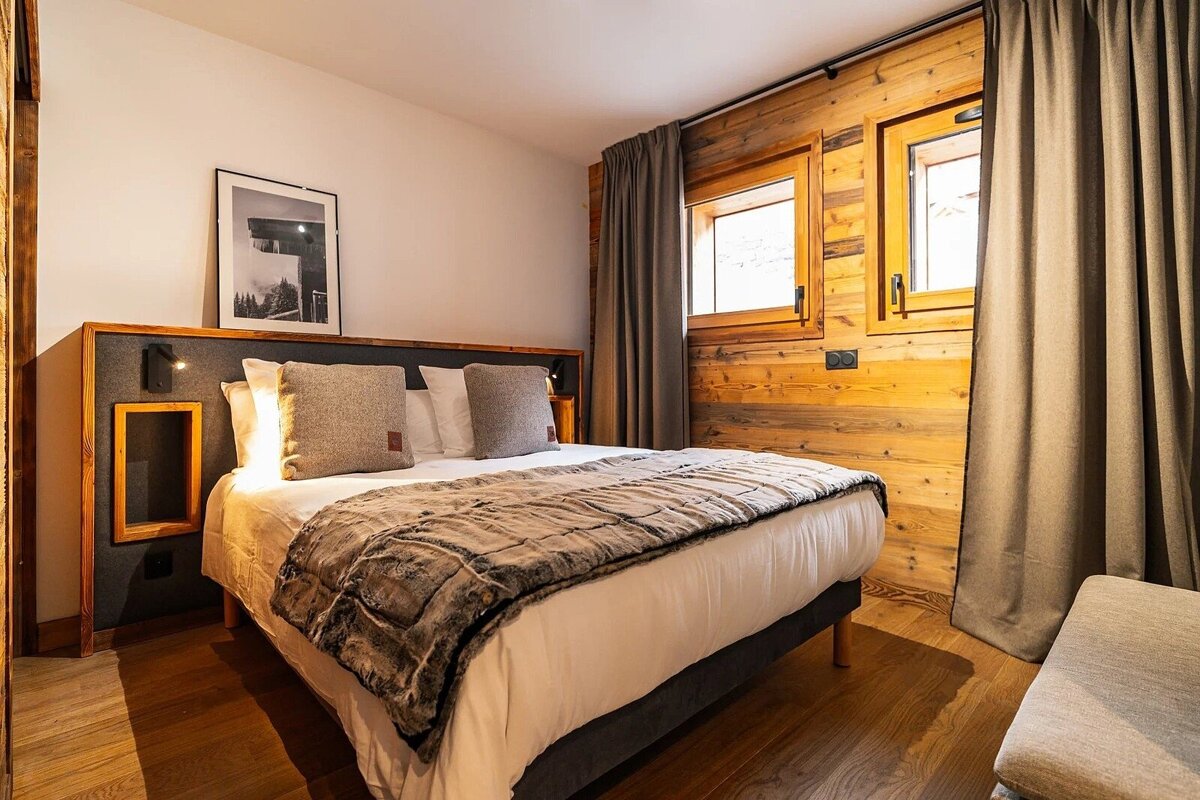 Cozy rustic bedroom featuring a double bed with a textured blanket, wooden walls, grey curtains, and warm lighting.